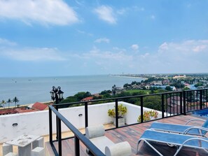Terraço no telhado com vista para o mar, um edifício branco e uma igreja ao longe.