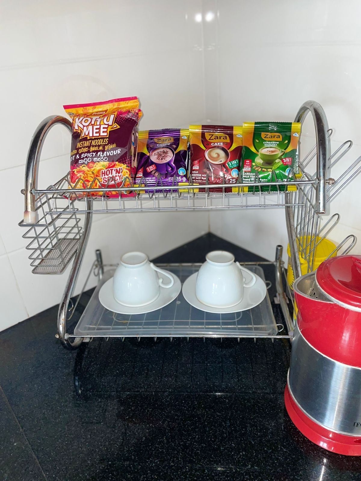 A kitchen area with a dish rack holding instant noodles, chai tea, and coffee.
