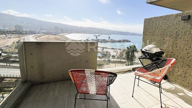 A balcony with two wicker chairs, a grill, and a view of a beach and cityscape.