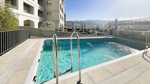 A swimming pool with metal ladders, surrounded by a tiled deck and a metal fence.