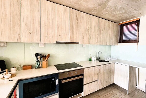 A modern kitchen with wooden cabinets, a built-in oven, and a large window with blinds. - Exclusivo y moderno depto. Mirador Barón (Valparaíso)