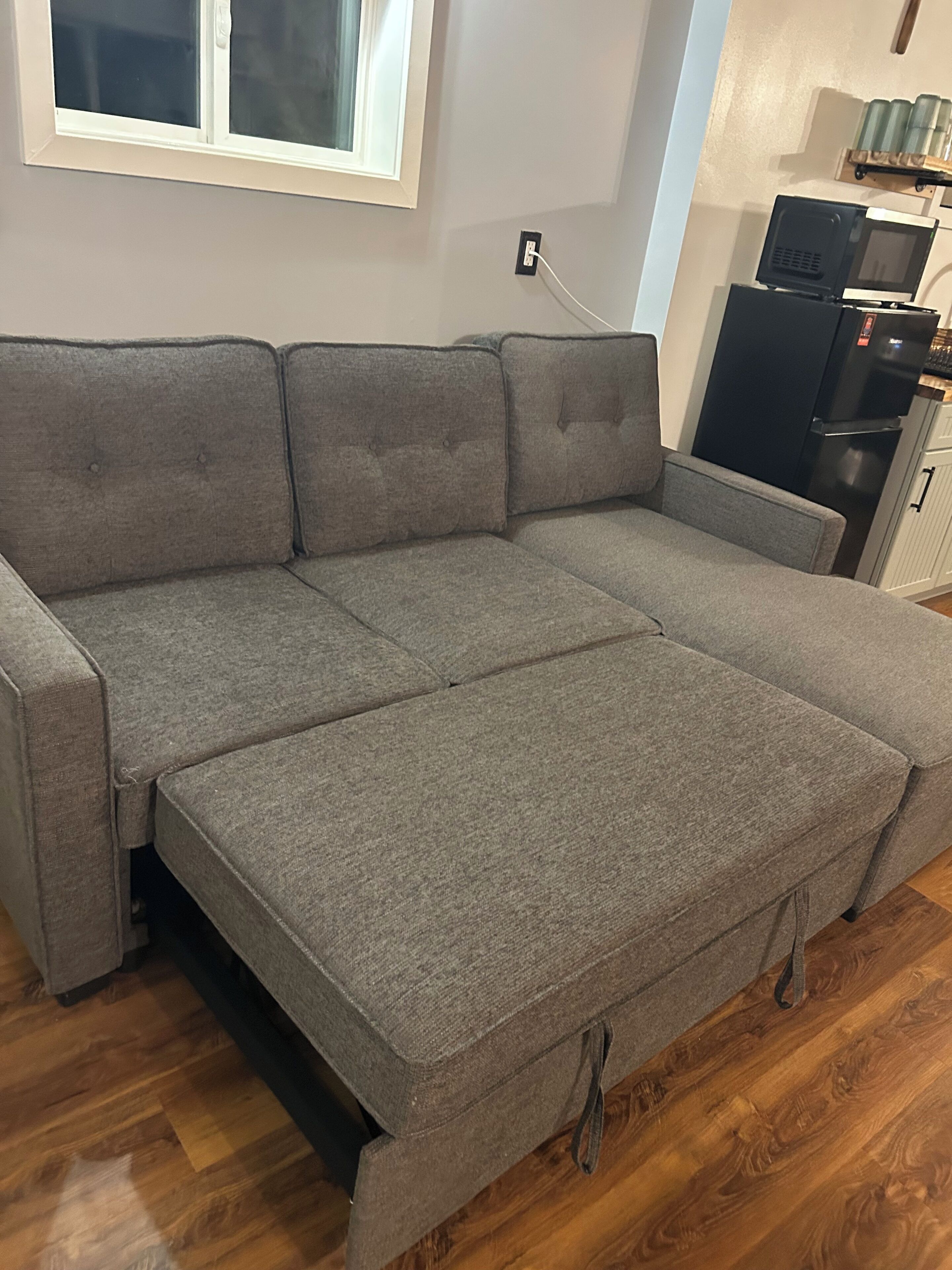 A grey sofa with a pull-out section, a window above it, and a kitchen area with a microwave and refrigerator in the background.