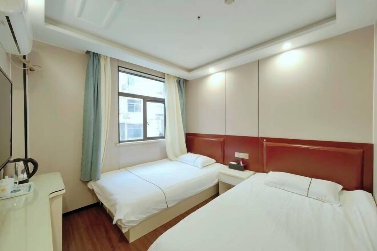Een hotelkamer met twee enkele bedden, een nachtkastje met een lamp, een klein bureau met een telefoon en een raam met gordijnen.