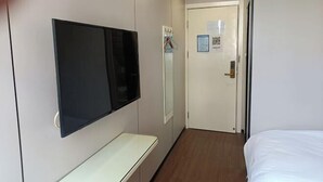 Una habitación de hotel con una cama, un televisor de pantalla plana montado en la pared, una mesita de noche y una puerta con espejo y perchero.