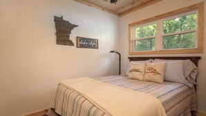 A bedroom with a bed, a window with a view of trees, a wall-mounted lamp, and a decorative sign.