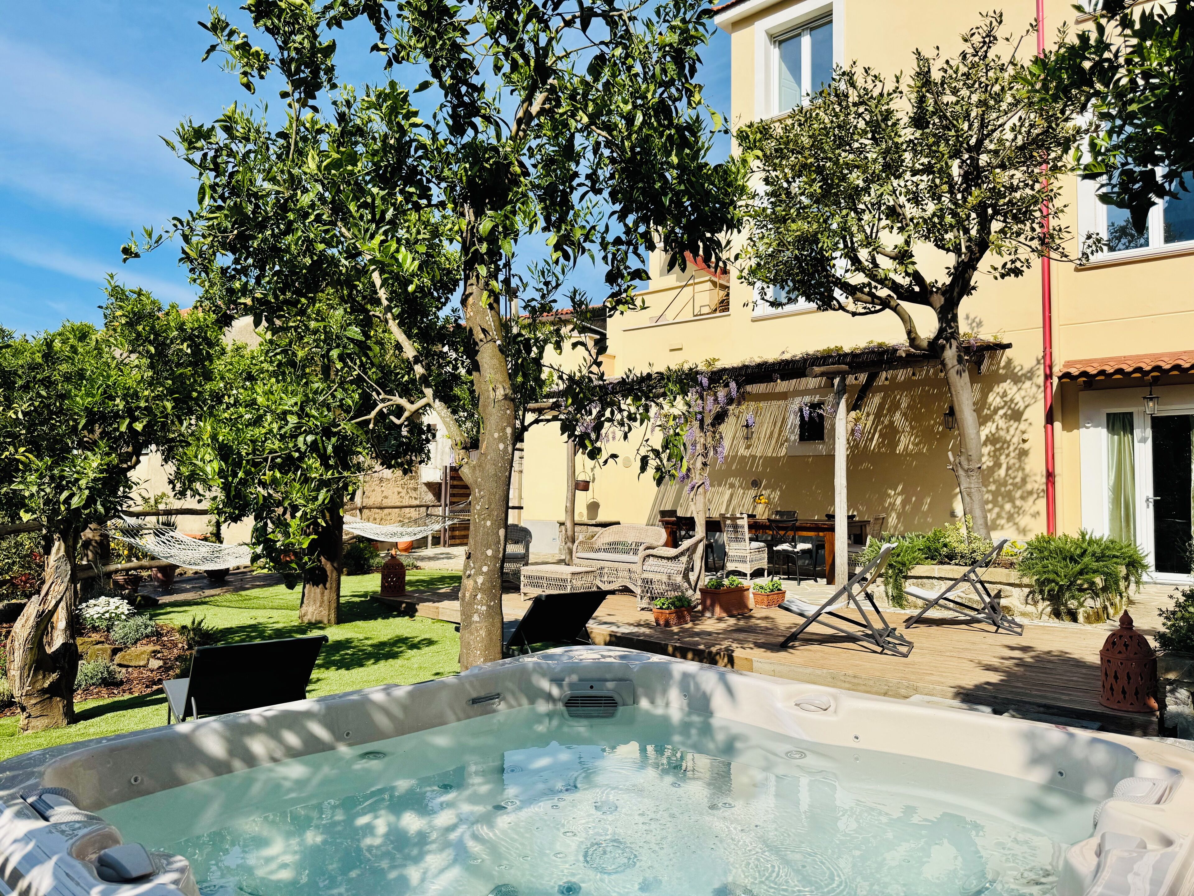 Un bain à remous dans un espace extérieur ensoleillé, agrémenté d’une pergola, de chaises longues et d’un bâtiment en arrière-plan.