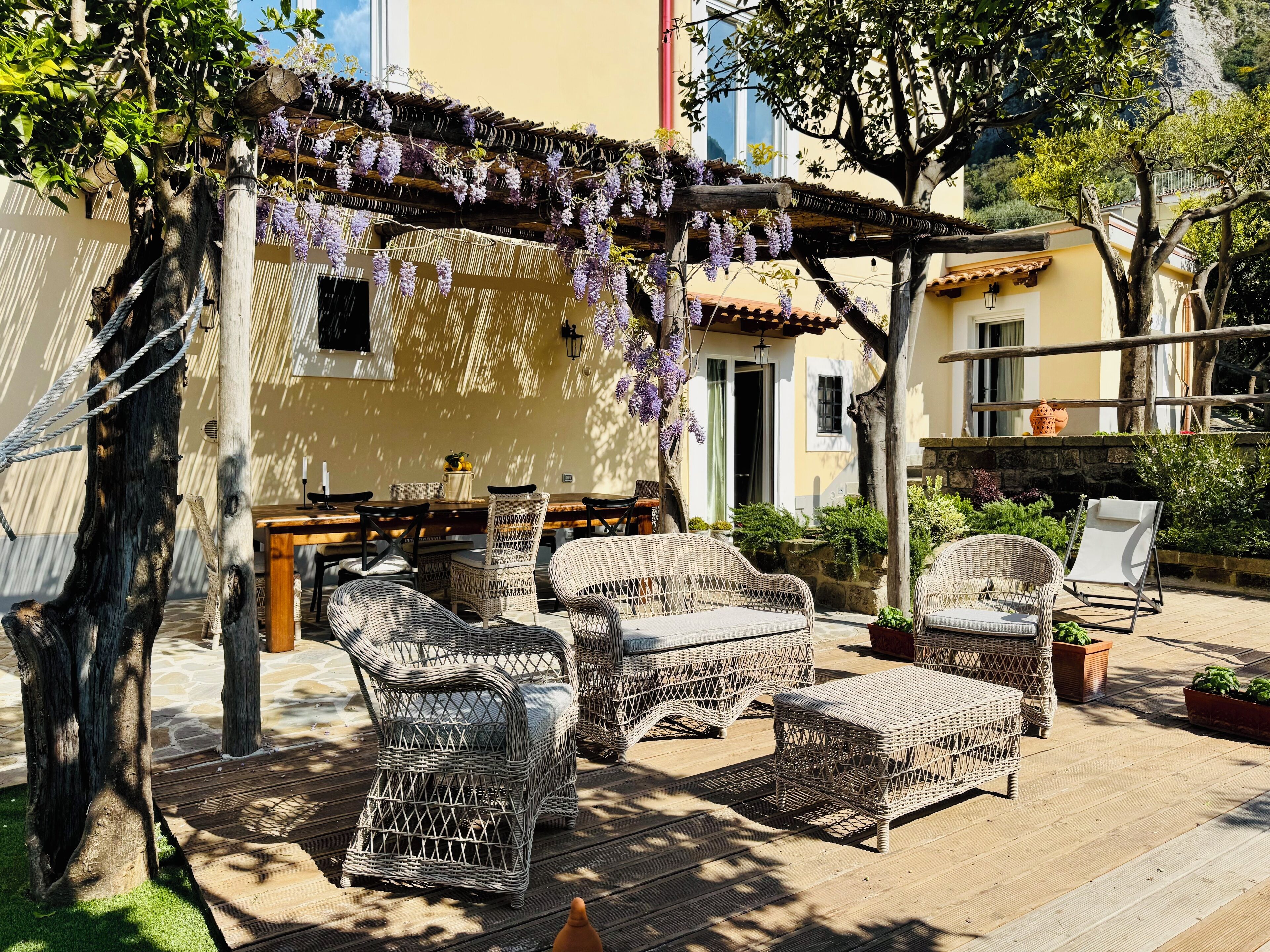 A patio with wicker furniture, a pergola with hanging purple flowers, and a stone wall.