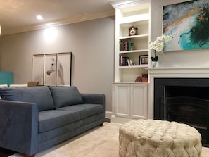 A living room with a grey sofa, a white ottoman, a fireplace, and a bookshelf with books and decorative items. - 5 Blocks to US Capitol, Renovated 1895 Capitol Hill Victorian (Washington)