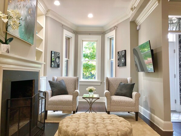 A living room with two patterned armchairs, a beige ottoman, a fireplace, and a large window with a view of greenery.