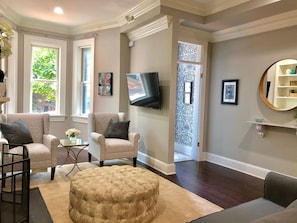 A living room with two armchairs, a round ottoman, a flat-screen TV, a patterned wall, and a bookshelf. - 5 Blocks to US Capitol, Renovated 1895 Capitol Hill Victorian (Washington)
