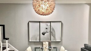 Uma sala de jantar com mesa de vidro, cadeiras brancas, lustre e uma pintura de um cavalo emoldurada na parede.