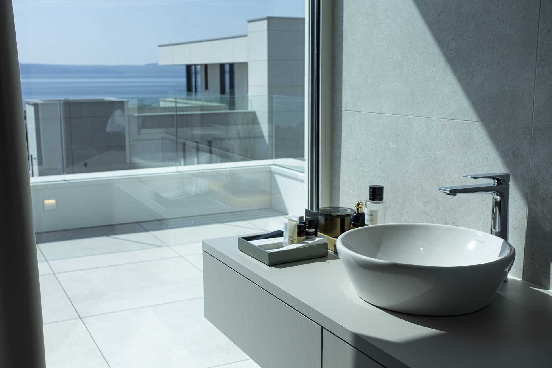 A modern bathroom with a large white sink, a glass shower enclosure, and a view of the ocean.