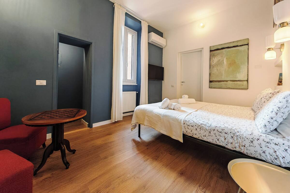A room with a bed, a small table, a red chair, and a window with curtains.