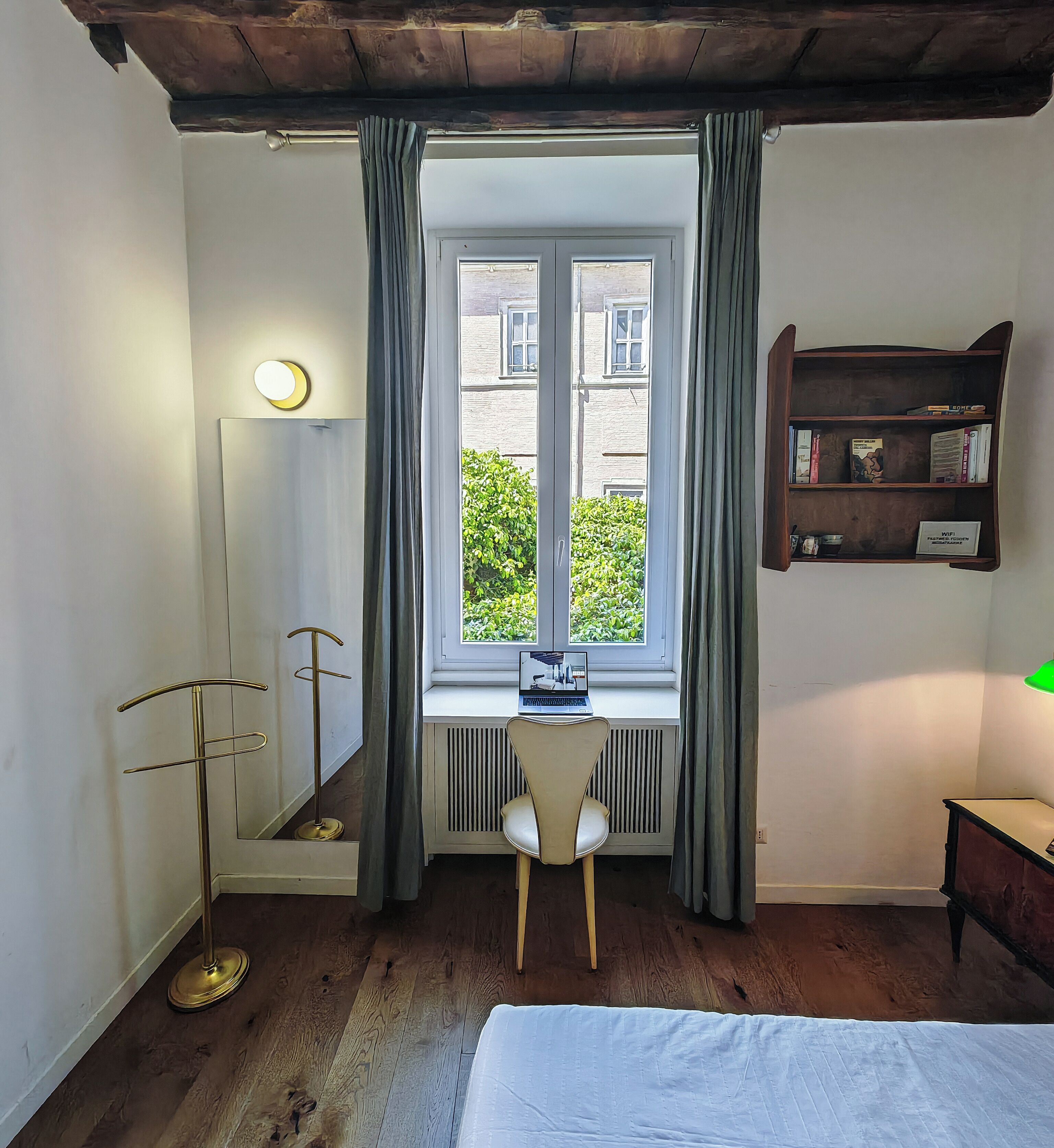 Una habitación con una cama, un escritorio con una computadora portátil, una lámpara de pie, una estantería y un ventanal con cortinas.