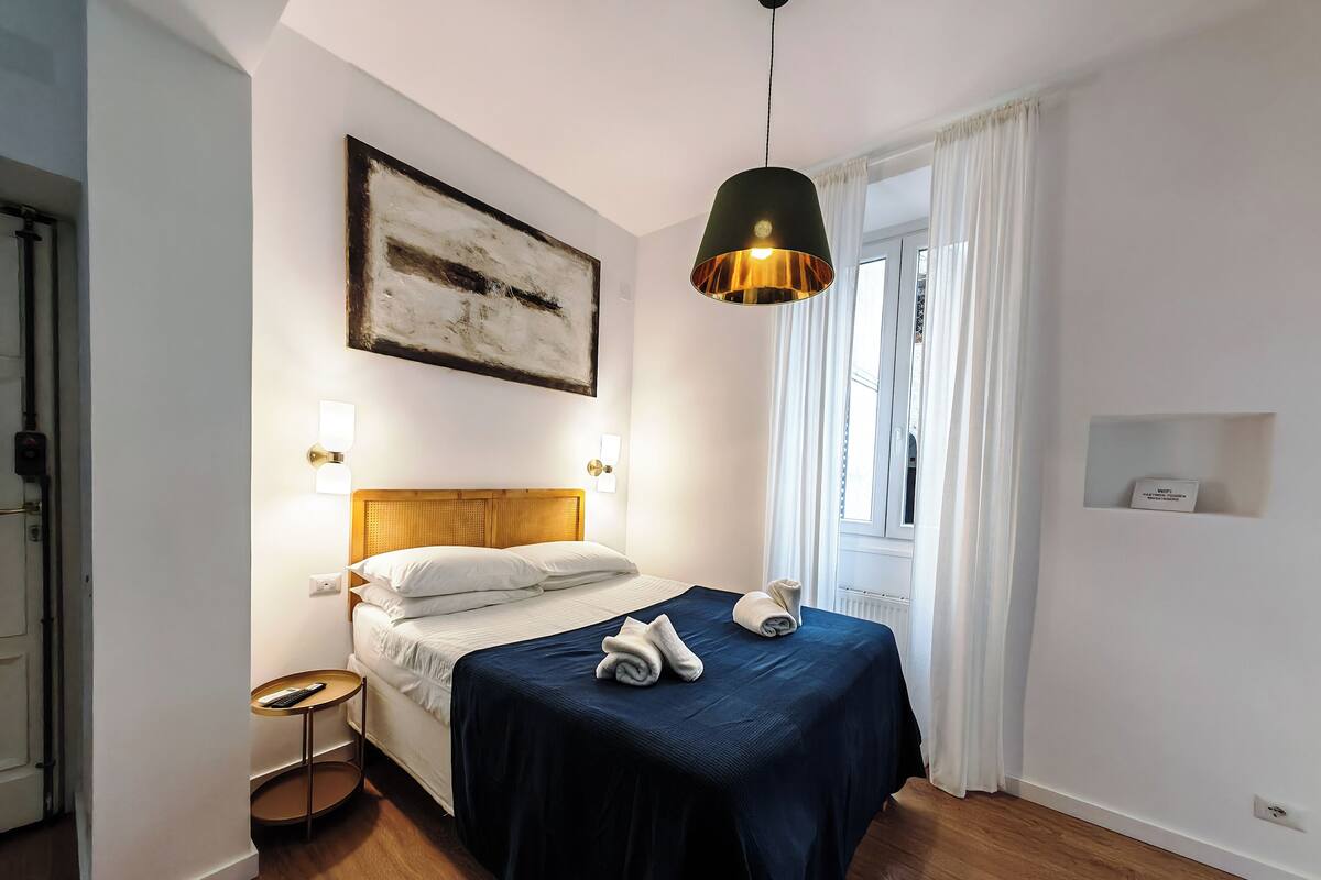 A bedroom with a wooden headboard, a bed with a dark blue bedspread, a hanging pendant light, and a painting on the wall.