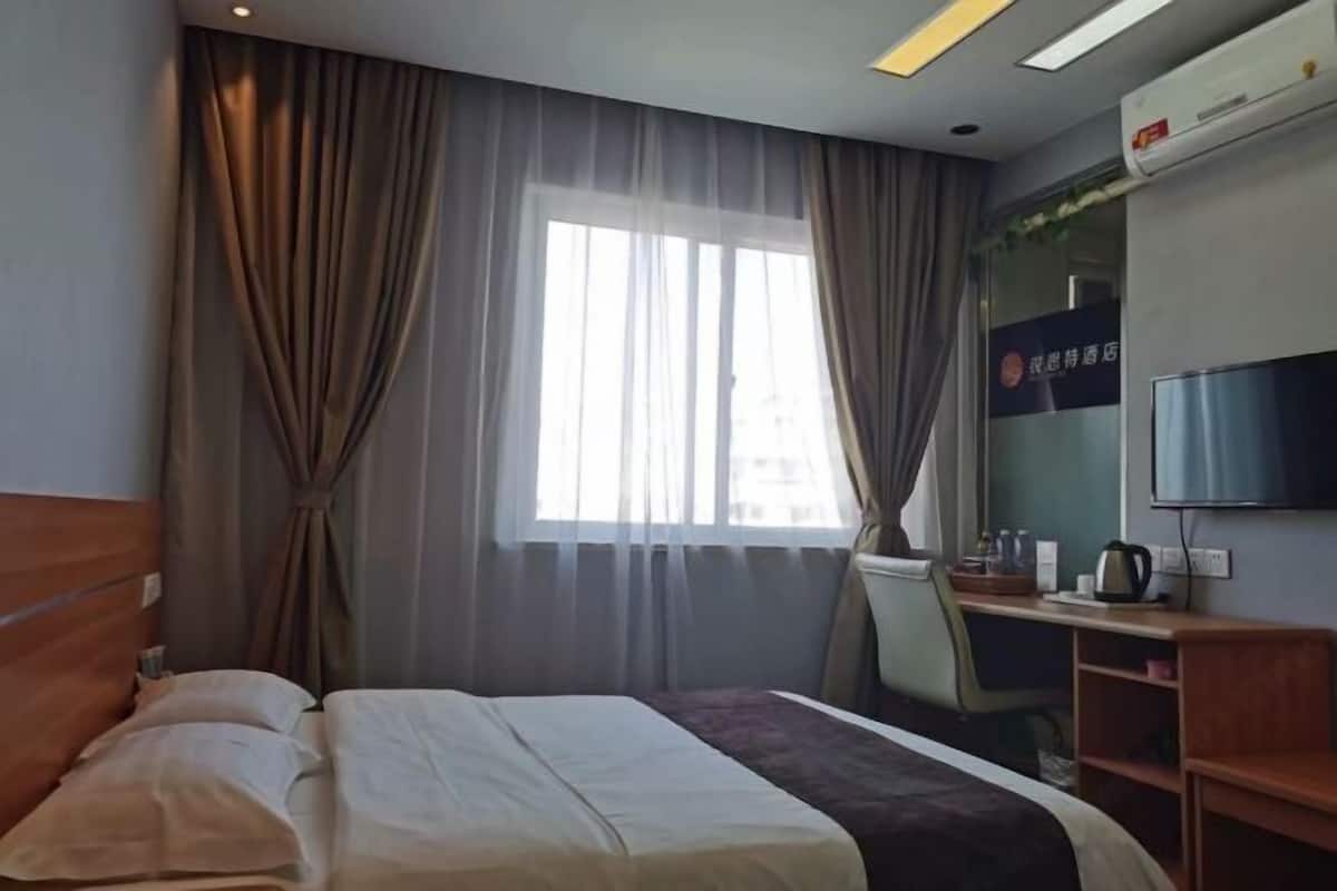 A hotel room with a bed, curtains, a desk with a chair, a television, and an air conditioning unit.
