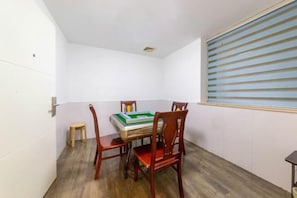 Um quarto pequeno com soalho de madeira, uma mesa com cadeiras, uma janela com persianas e um banquinho.