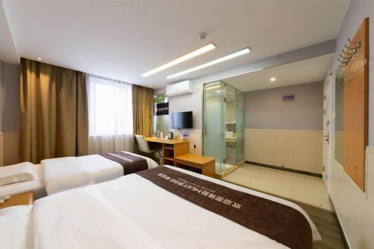 A hotel room with two beds, a desk with a chair, a television, and a glass door leading to a bathroom.