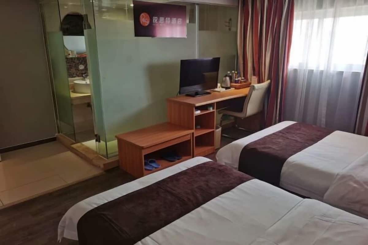 A hotel room with two beds, a desk, a chair, a TV, and a bathroom visible through a glass partition.