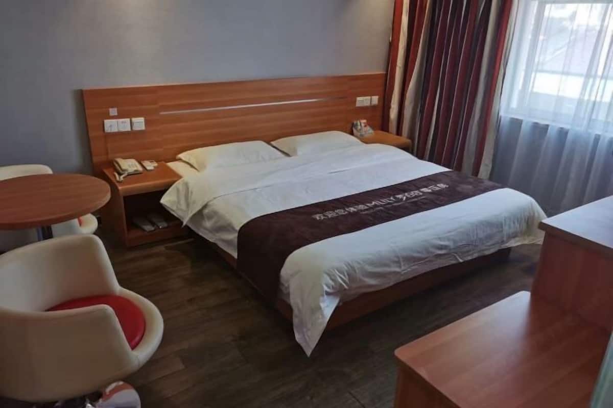 A hotel room with a wooden headboard, a bed with a white and brown bedspread, a small bedside table with a phone, a chair with a red cushion, and a wooden desk.