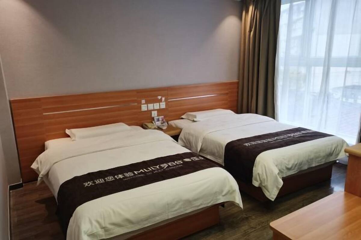 A hotel room with two beds, a wooden headboard, a bedside table, and a window with curtains.