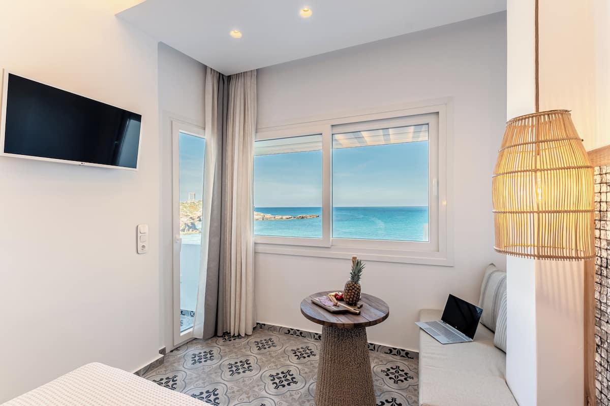 A modern hotel room with a large window offering a sea view, a flat-screen TV mounted on the wall, a laptop on a sofa, and a small round table with a pineapple and a tray of fruit.