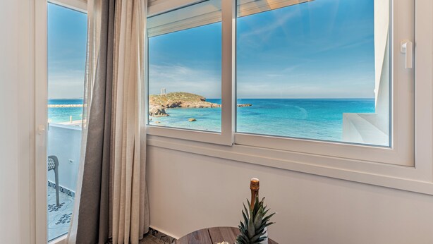 Una habitación con vistas al mar a través de amplios ventanales, una cortina y una pequeña mesa con una piña.
