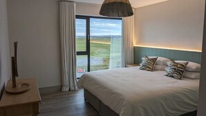 A bedroom with a large bed, a wooden nightstand, a hanging lamp, and a view of a green landscape through a glass door.