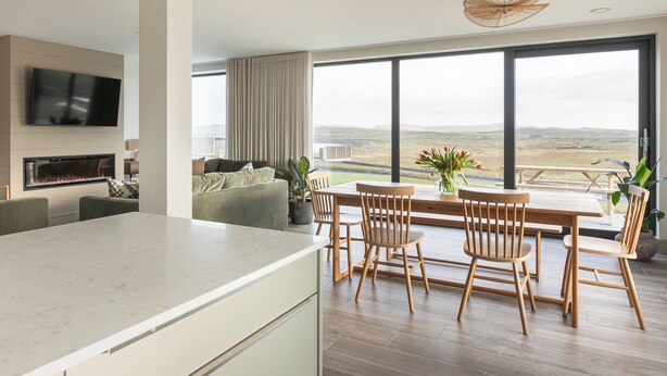 A modern kitchen with a large island, a dining area with wooden chairs, a flat-screen TV mounted above the kitchen, and a large window offering a view of the countryside.