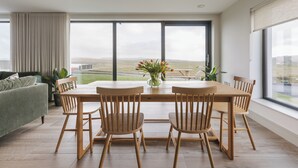 A modern dining area with a wooden table and chairs, large windows with a view of the countryside, and a green sofa in the background.