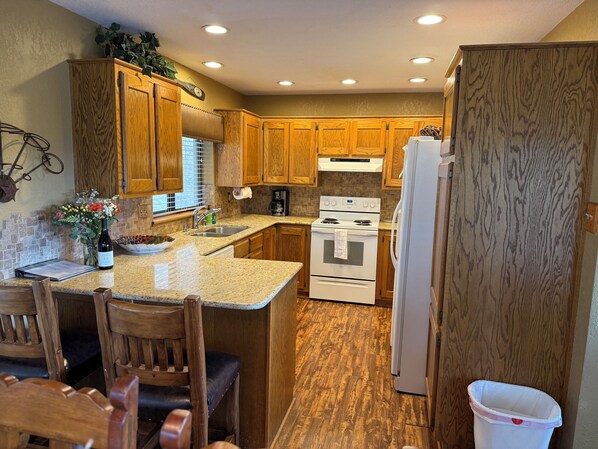 A kitchen with wooden cabinets, a granite countertop, a stove, a refrigerator, and a dining area with chairs.