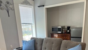 Un sofá seccional gris con cojines azules en una sala de estar con piso de madera, una ventana con persianas, un televisor de pantalla plana y un refrigerador.