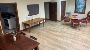 A spacious dining area with a wooden table and chairs, a kitchen in the background, and a large window providing natural light.