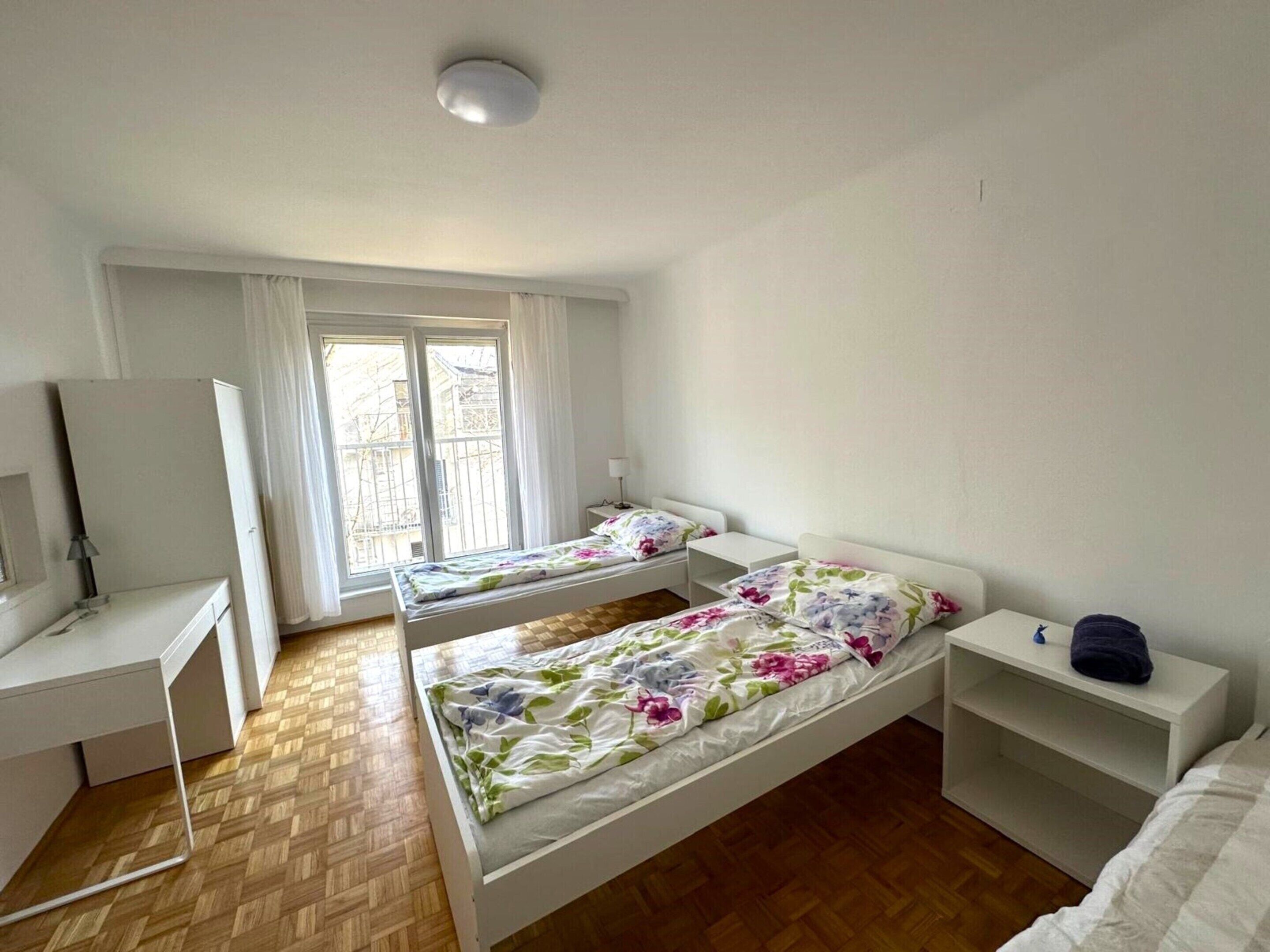 A small, bright room with two single beds, a desk, and a window with curtains.