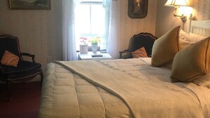 Ein Hotelzimmer mit einem großen Bett, zwei Sesseln, einem kleinen Tisch mit Blumenvase und einem Fenster mit Vorhängen.