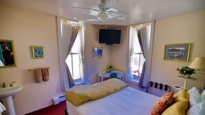 A hotel room with a bed, a desk, a chair, a TV, a window with curtains, a radiator, and a sink.
