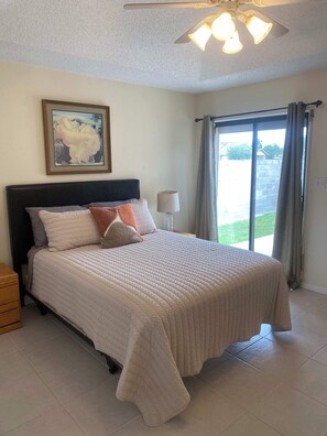A bedroom with a large bed, a ceiling fan, a painting on the wall, a nightstand, a lamp, and a window with curtains.