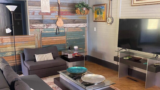A modern living room with a sectional sofa, a glass coffee table, a flat-screen TV, and a rustic wall with a guitar and plants. - #1 Guest House 2Bd 2 Bath Pet Friendly Across from Spa (Silverton)