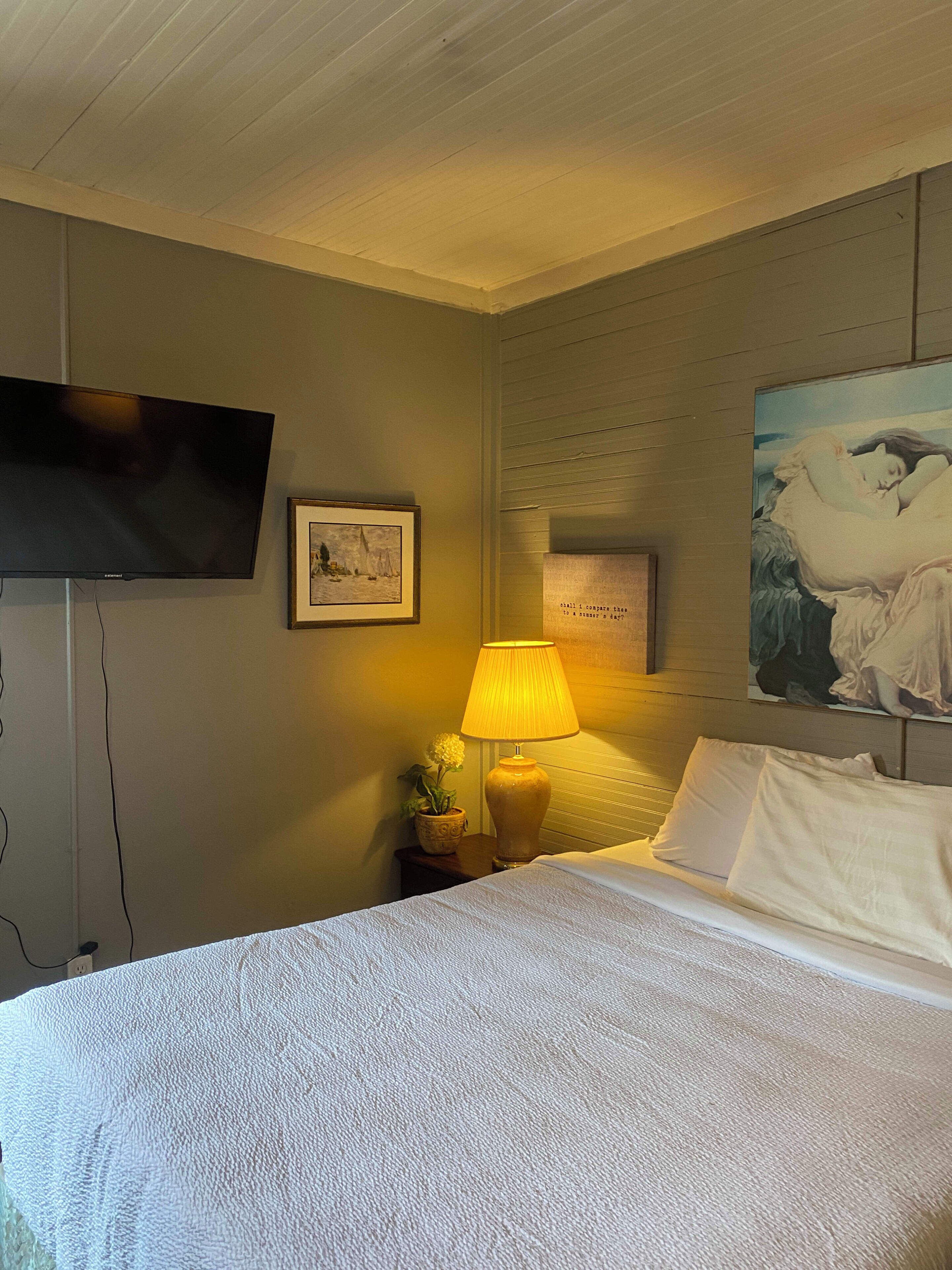 Ein Hotelzimmer mit einem Bett, einem an der Wand befestigten Fernseher, einem Gemälde, einer Lampe und einem kleinen Tisch mit einer Blume.