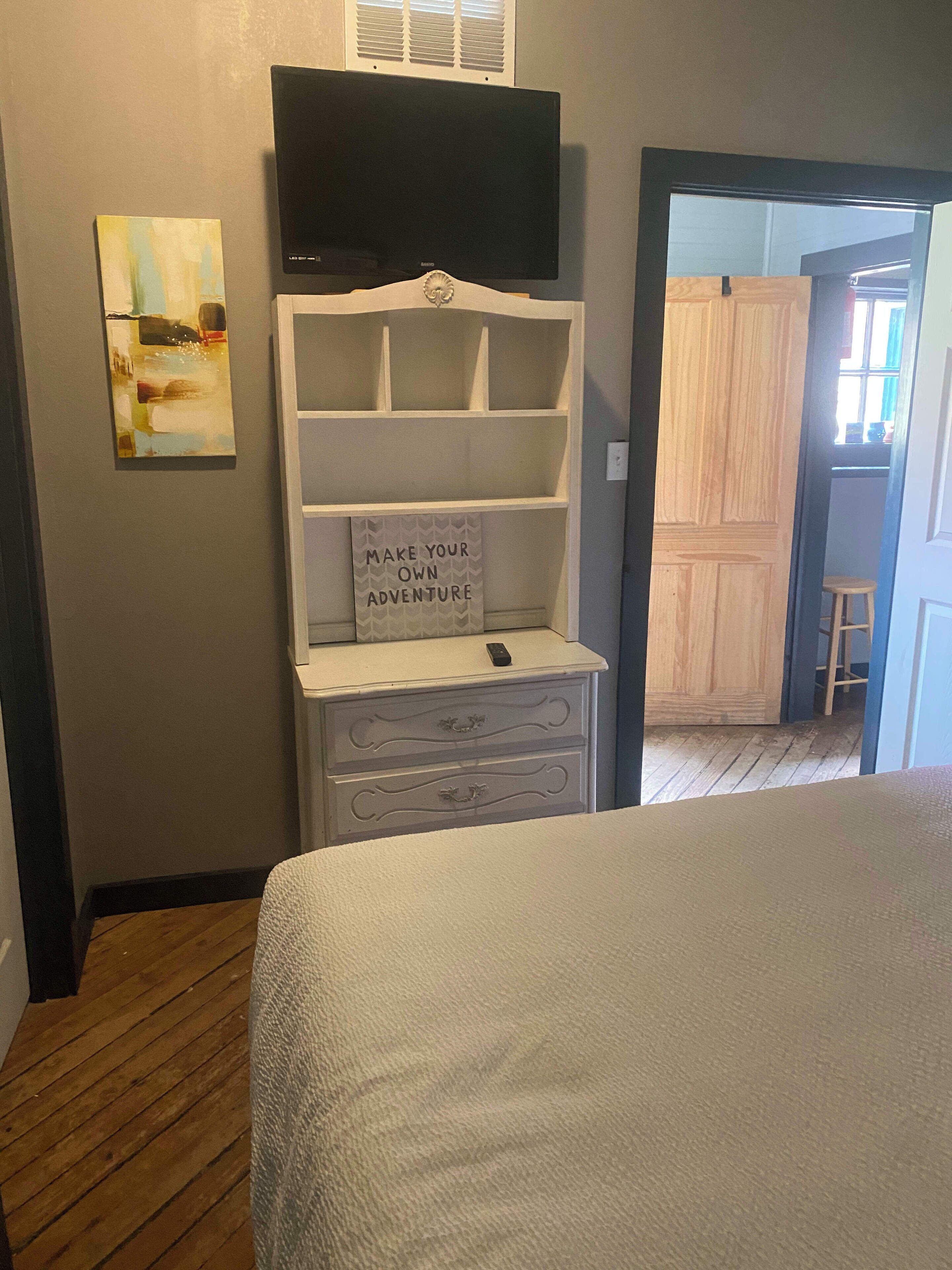A bedroom with a bed, a TV on a stand, a shelf with a motivational sign, and a wooden door leading to another room.