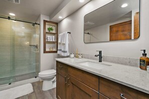 A modern bathroom with a glass-enclosed shower, a white toilet, a large mirror, and a marble countertop with a soap dispenser. - #1Condo Glacier Resort, Pool, Golf, HotTub, Sauna, Gym (Durango)