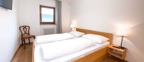 Un lit double avec une tête de lit en bois, deux tables de chevet avec des lampes, une chaise, une fenêtre donnant sur un beau paysage et un téléviseur fixé au mur.