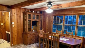 A wooden dining room with a large table and chairs, a ceiling fan, and a window with a view of trees.