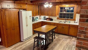 A kitchen with wooden cabinets, a white refrigerator, a wooden table with two stools, and a window with a view.