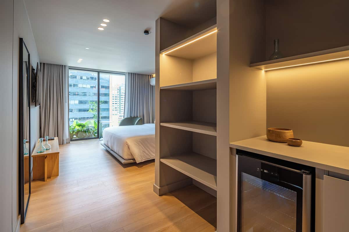 Una habitación de hotel moderna con una cama grande, un armario empotrado, una pequeña zona de cocina y vistas a la ciudad a través de amplias ventanas.
