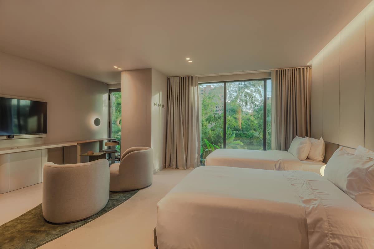 Una habitación de hotel moderna con dos camas, un televisor de pantalla plana, una zona de estar y ventanas amplias con cortinas.