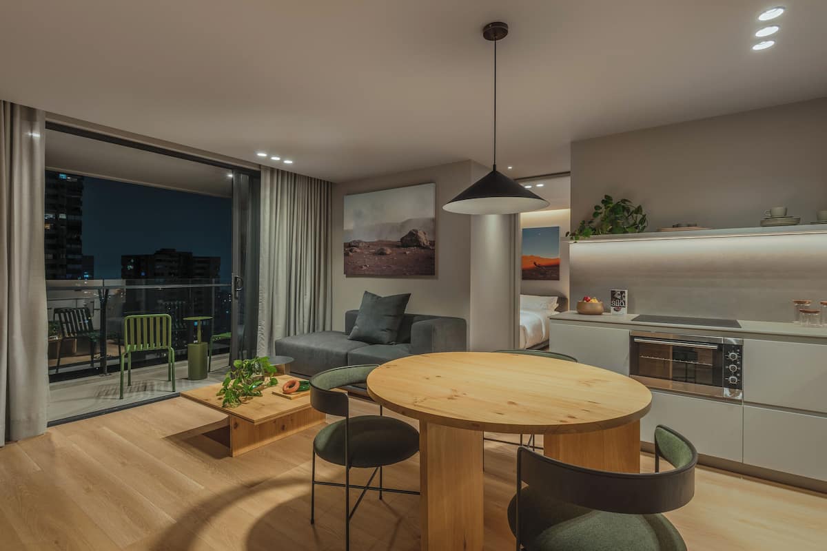 Una sala de estar moderna con una mesa de comedor de madera, sillas verdes, un sofá gris y un ventanal con vistas a un paisaje urbano.