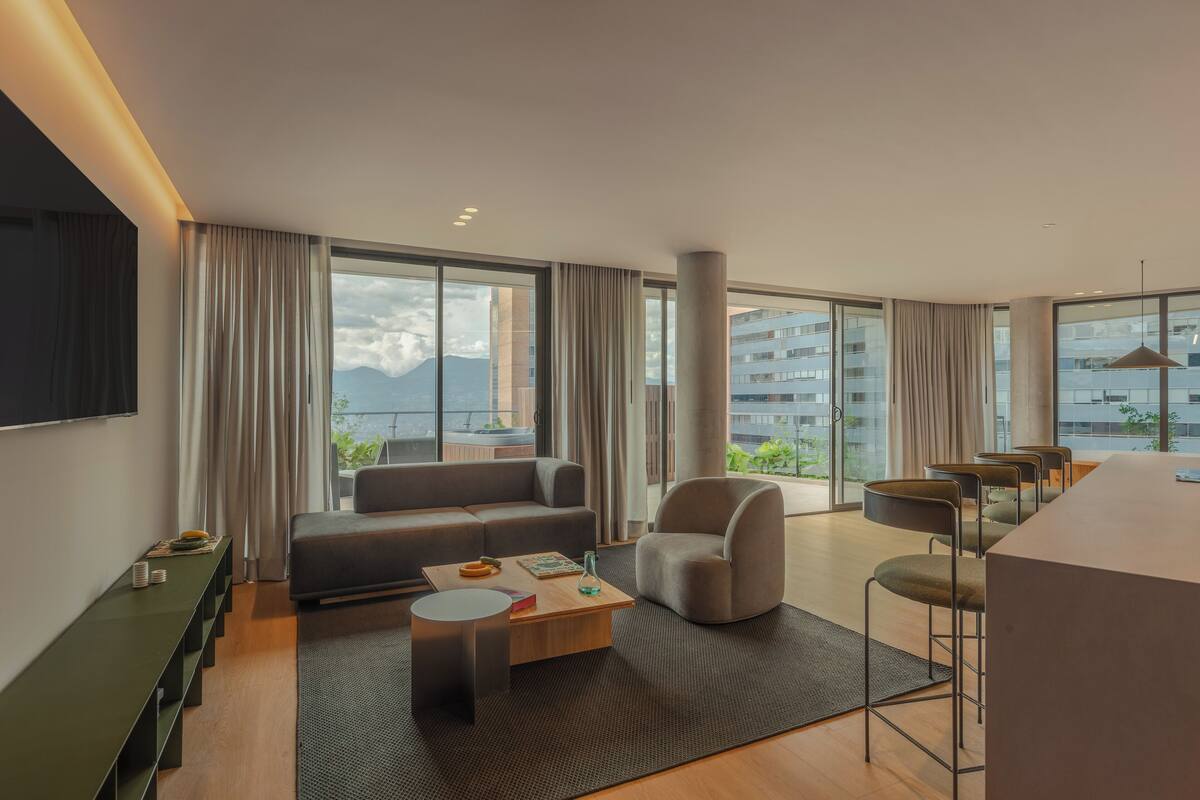 Habitación de hotel moderna con sofá, sillón, mesa de comedor y sillas. Grandes ventanales ofrecen vistas a la ciudad y a las montañas.