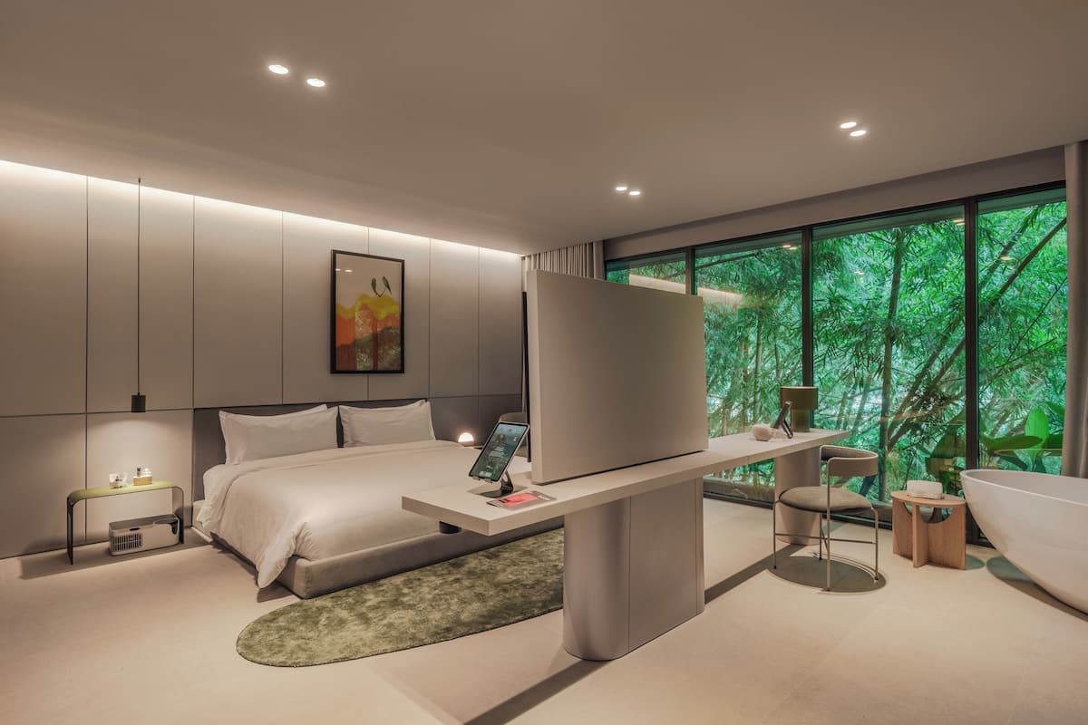 Un dormitorio moderno con cama, mesita de noche, televisor y un ventanal con vistas a árboles.