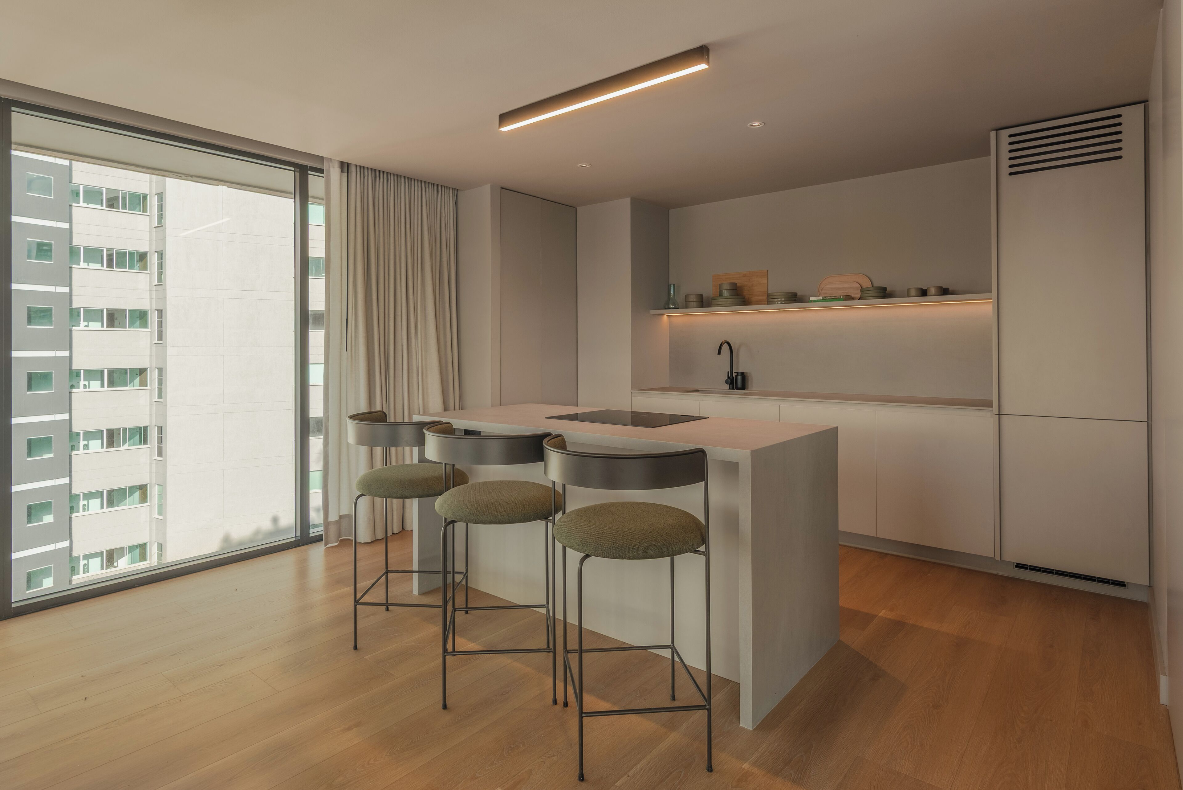 Uma cozinha moderna com uma ilha central, banquetas, uma janela ampla e vista para um edifício.